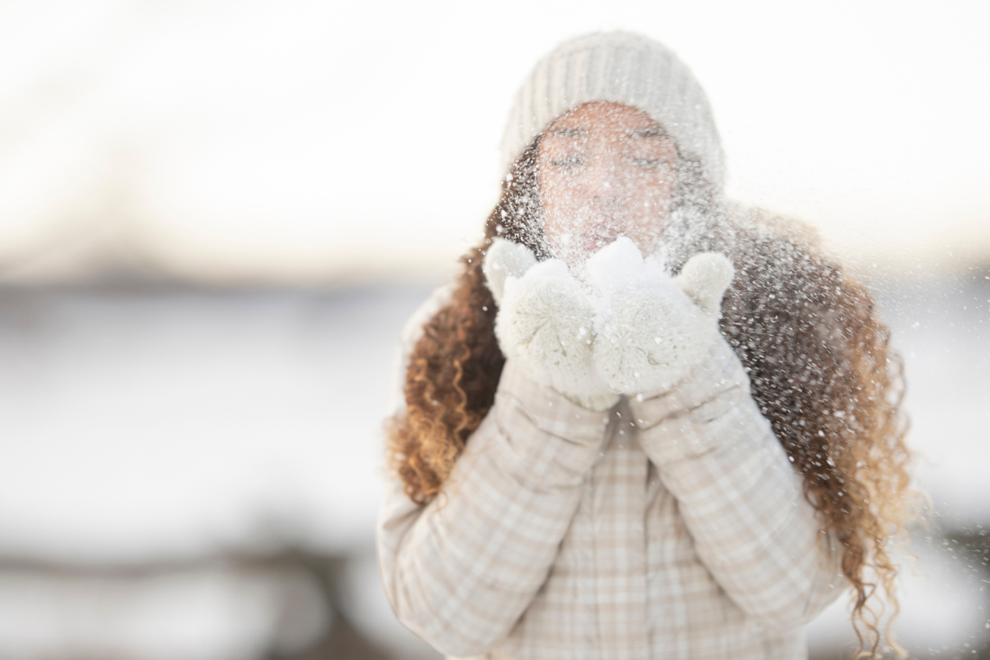 A woman with long, curly hair wearing winter clothing stands outdoors in the snow and blows snowflakes from her gloved hands. The image captures a soft, cozy winter moment and highlights natural textured hair in cold weather conditions.