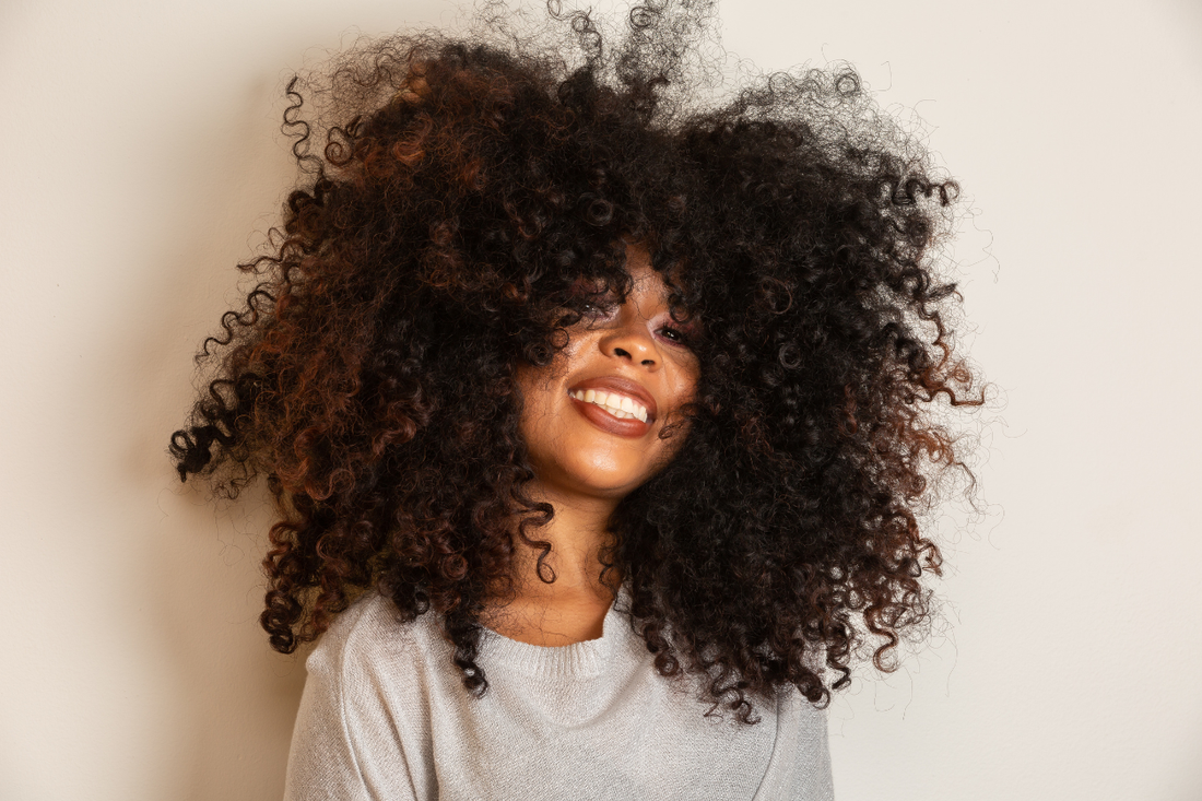 Woman with voluminous natural curls and defined texture smiling against a neutral background, showcasing healthy, full curly hair.”