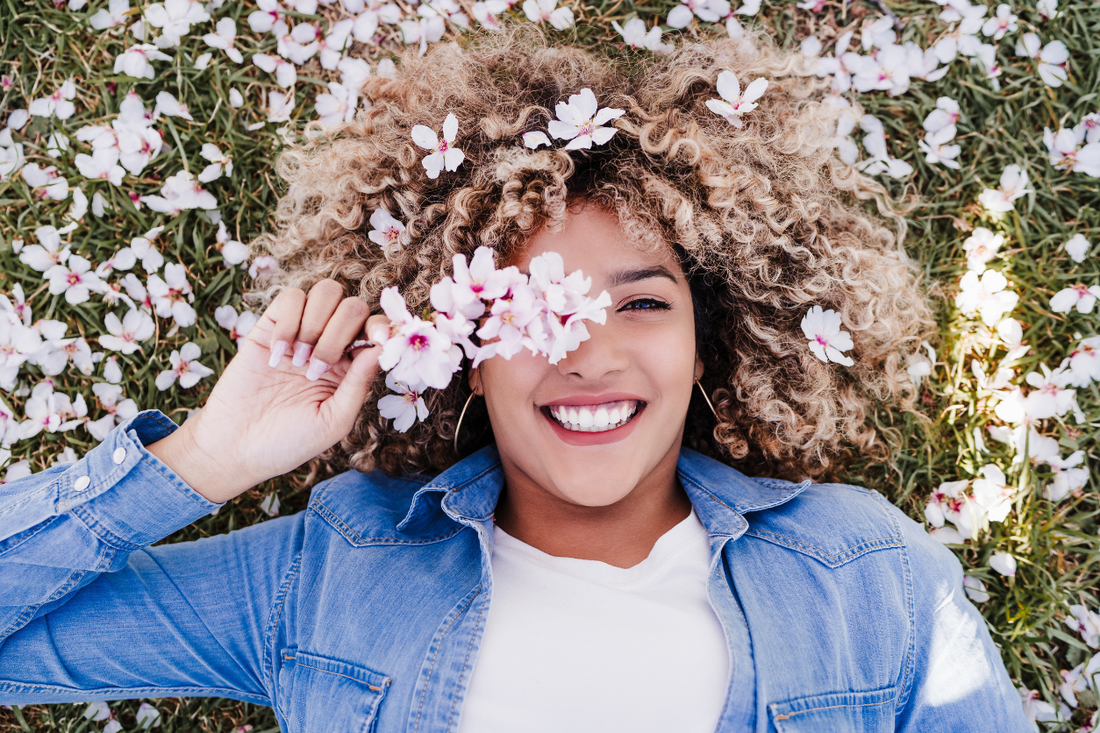A smiling woman with voluminous curly hair lying in a field of white flowers, holding a small cluster of blooms over one eye.