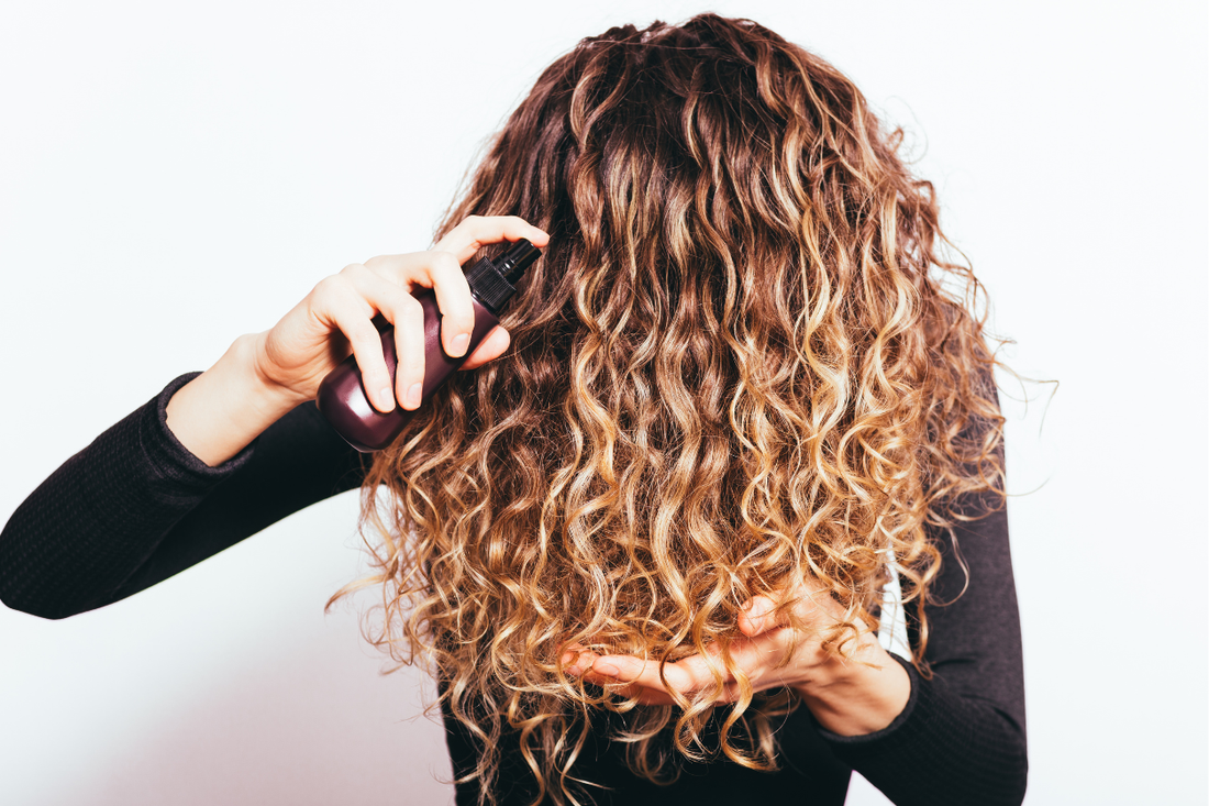 A woman with long, highlighted curly hair misting a spray bottle through her curls against a white background.