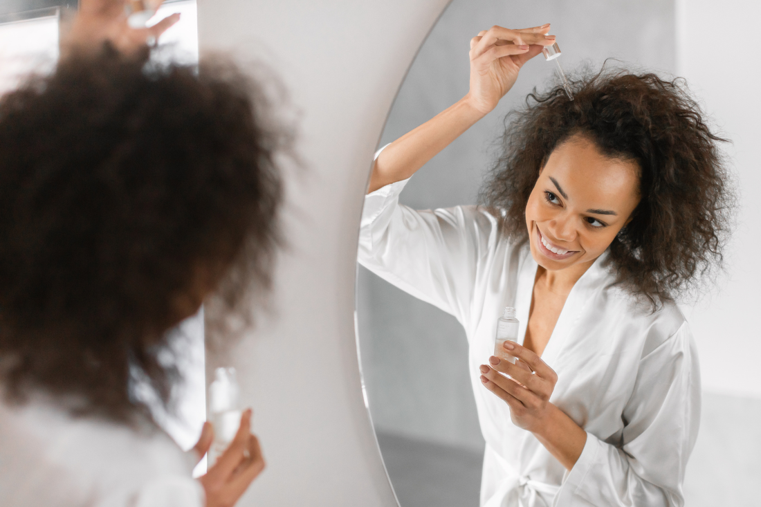 Woman applying a hair serum with a dropper to her curls while facing a round mirror
