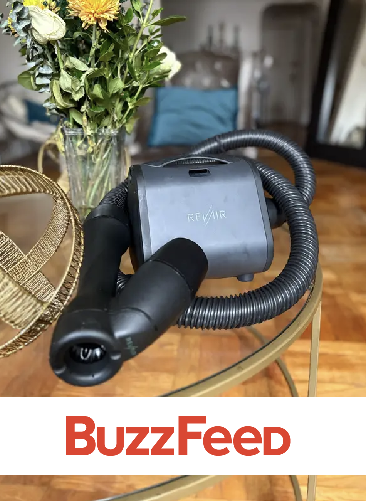 RevAir Reverse-Air Dryer unit and wand resting on a glass side table next to a vase of flowers in a living room setting, with the BuzzFeed logo displayed below