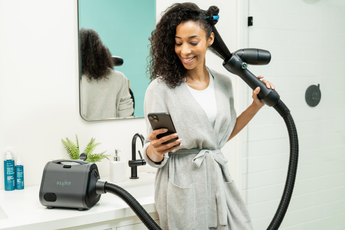 Woman with curly hair using a RevAir reverse-air dryer in a bathroom, smiling while checking her phone as a section of hair is stretched and dried with the wand.