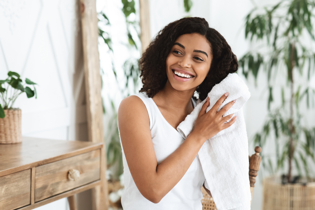 Smiling woman gently drying her curly hair with a soft white towel in a bright, plant-filled room