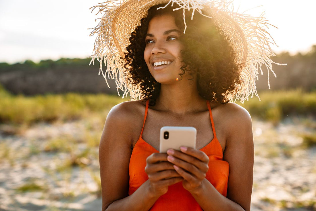 Woman with natural curly hair smiling on the beach while checking her phone in the summer sun.