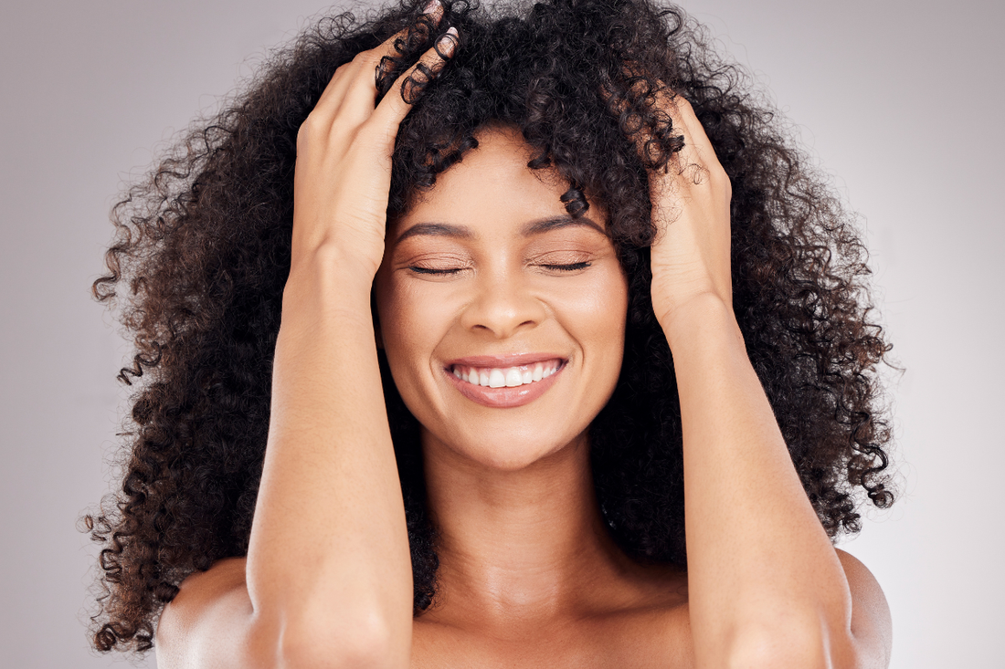 Close-up of a smiling woman with voluminous natural curls, eyes closed, hands placed in her hair, showcasing healthy textured hair against a soft neutral background.