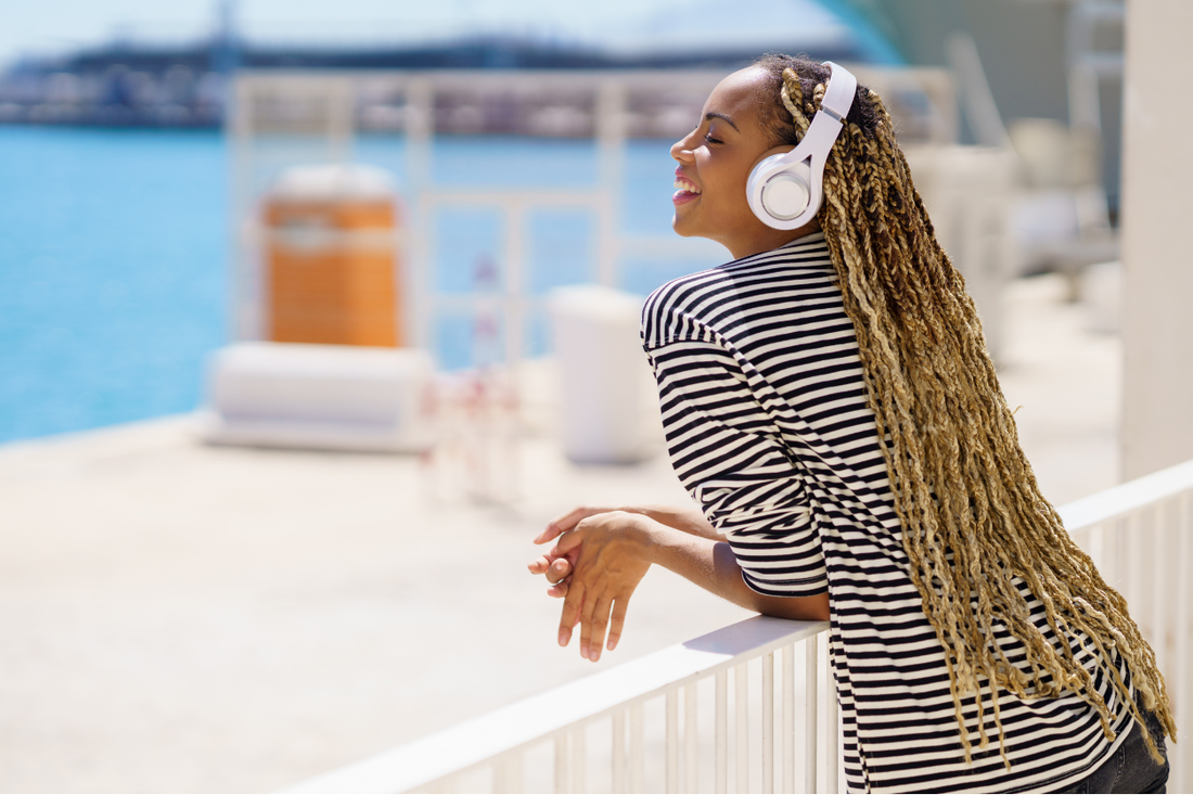 A Black woman with long blonde box braids wearing white headphones, leaning on a railing outdoors near the water on a sunny day.
