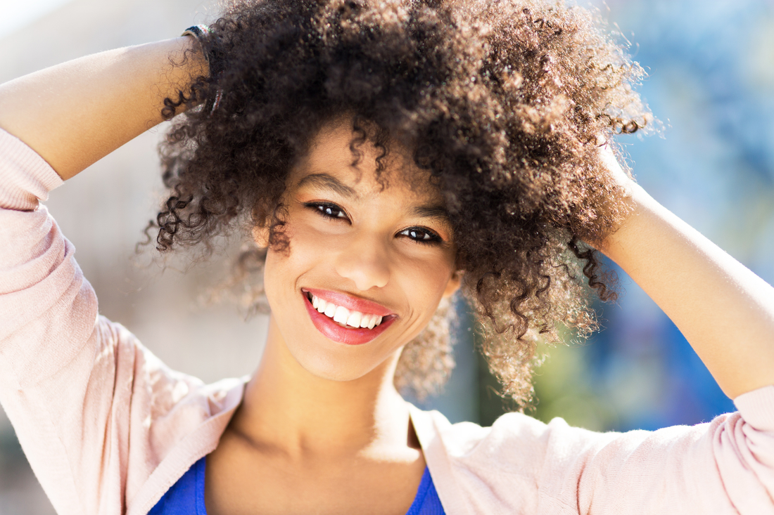 A smiling young woman with voluminous natural curly hair holding both hands up through her curls against a bright outdoor background.